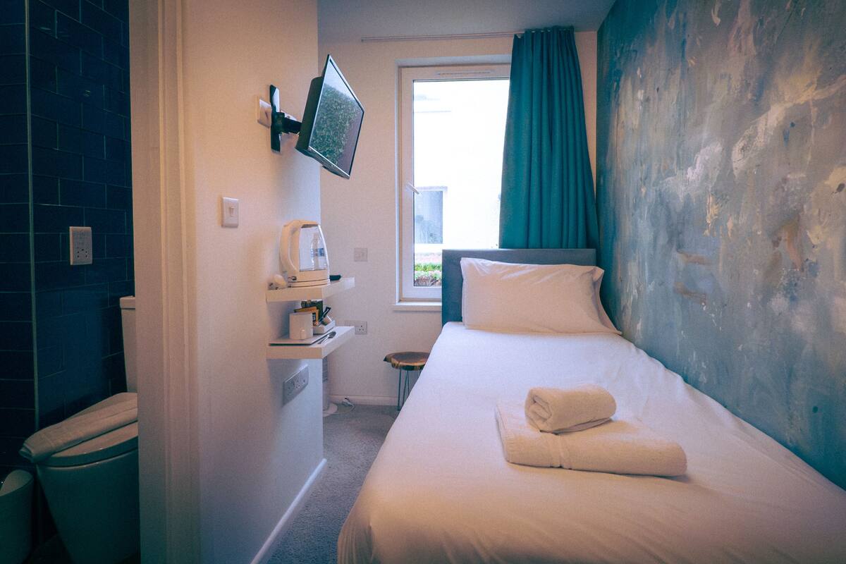 Ein Hotelzimmer mit Bett, einem an der Wand befestigten Fernseher, einem Fenster mit Vorhängen und Blick ins Freie.