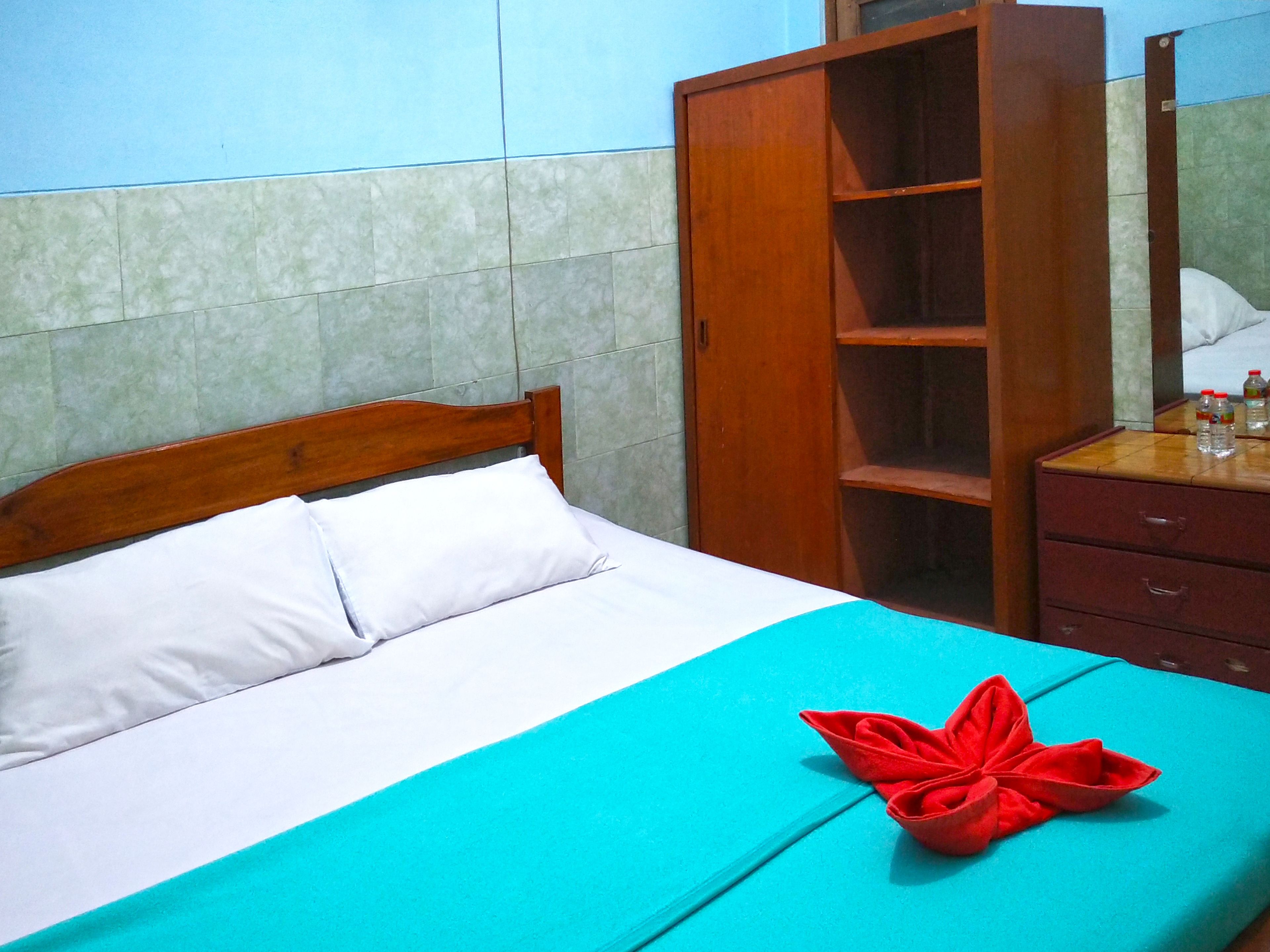 Una cama con almohadas blancas y una colcha azul turquesa, un armario de madera y una mesita con botellas de agua.
