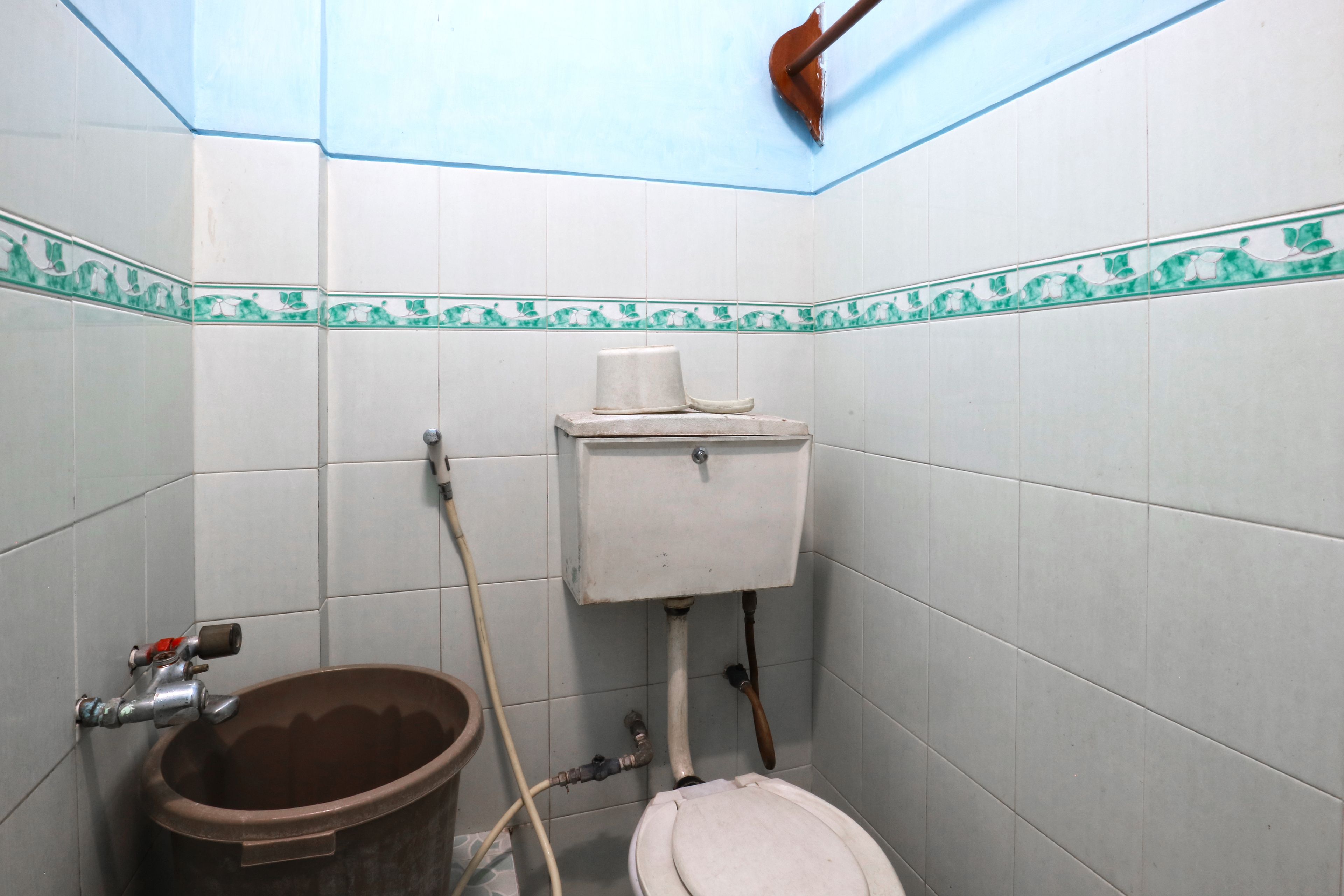 A bathroom with white tiled walls, a toilet, a brown bucket, and a visible plumbing system.