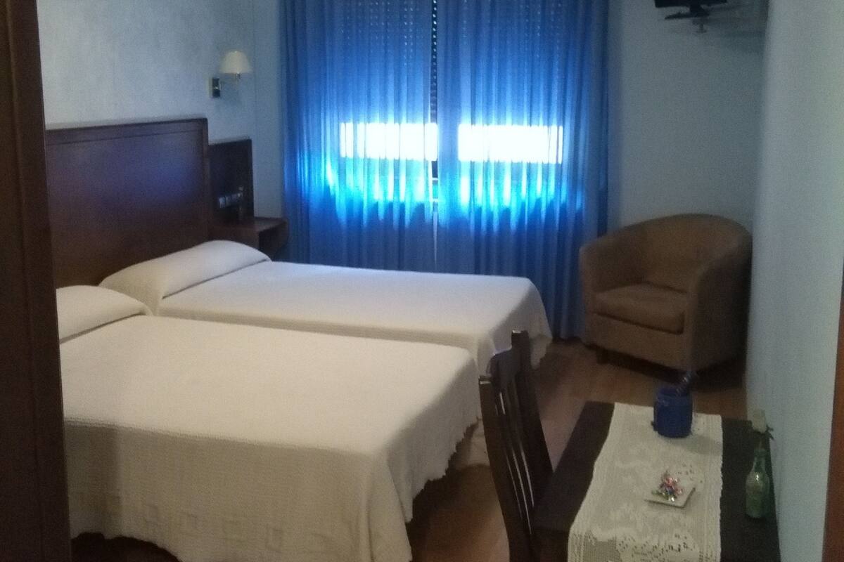 Quarto de hotel com duas camas, uma escrivaninha com cadeira, televisão na parede e janela com cortinas azuis.
