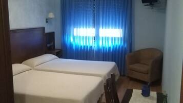 Habitación de hotel con dos camas, un escritorio con una silla, un televisor en la pared y una ventana con cortinas azules.