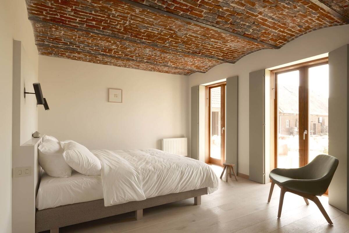 Een slaapkamer met een bakstenen plafond, een bed, een stoel en een raam met uitzicht op het omliggende landschap.
