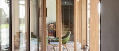 A rustic interior with a stone floor, wooden ceiling, and large glass doors leading to an outdoor area with a green chair and a small table.