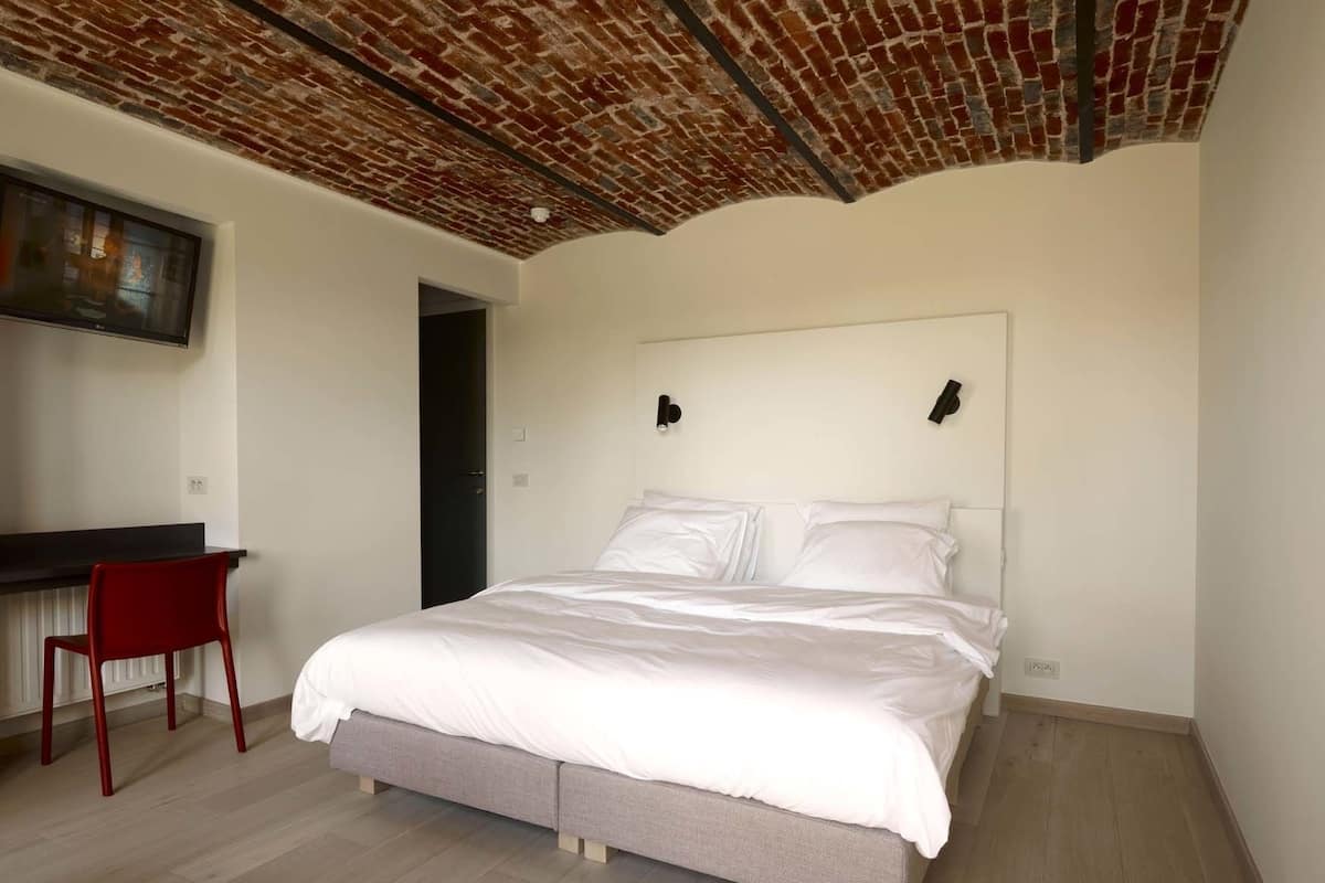 Een slaapkamer met een bakstenen plafond, een bed met wit beddengoed, een rode stoel en een klein bureau.