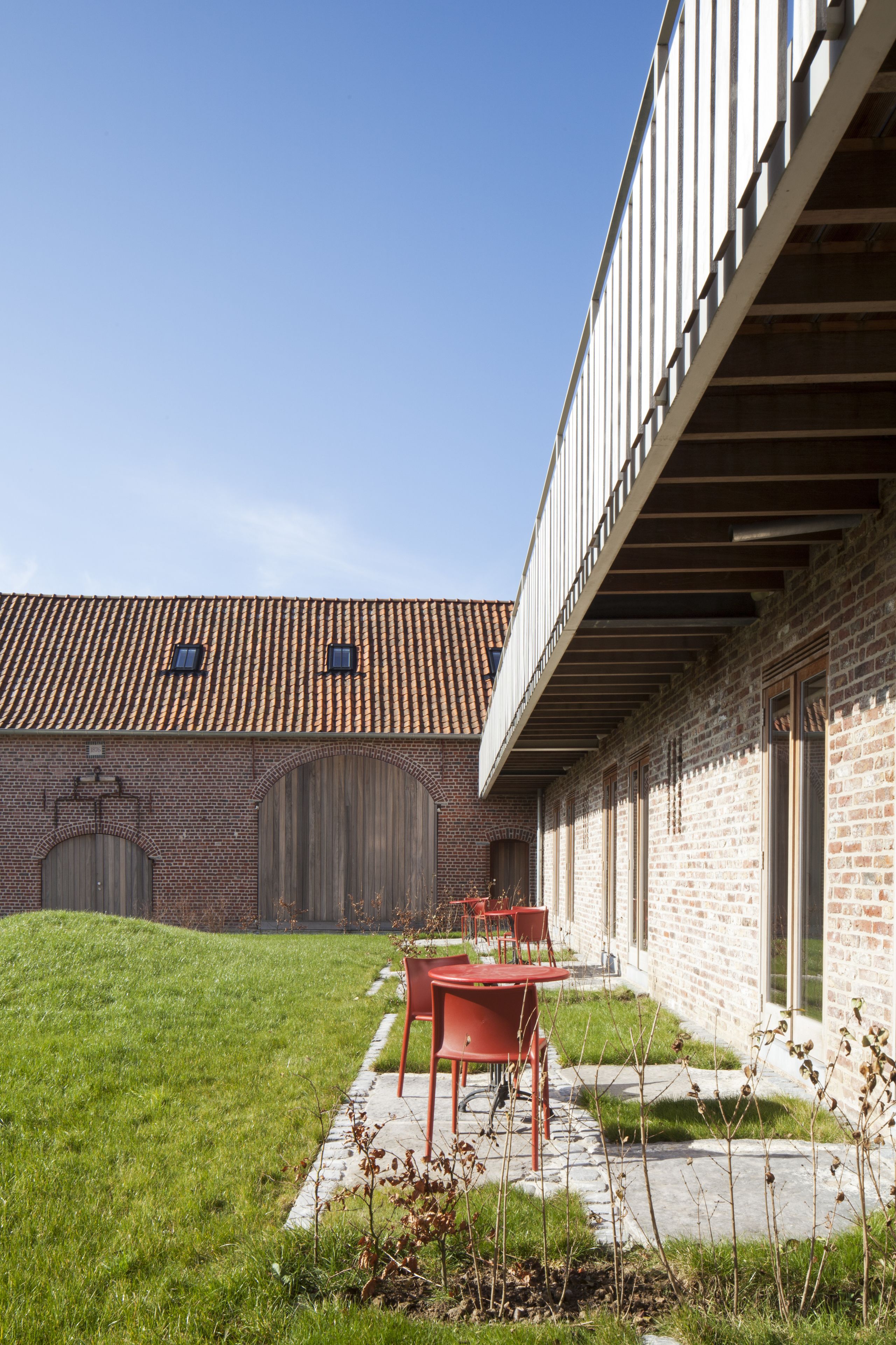 Une cour avec un bâtiment en briques, un toit recouvert de tuiles et une table et des chaises rouges.