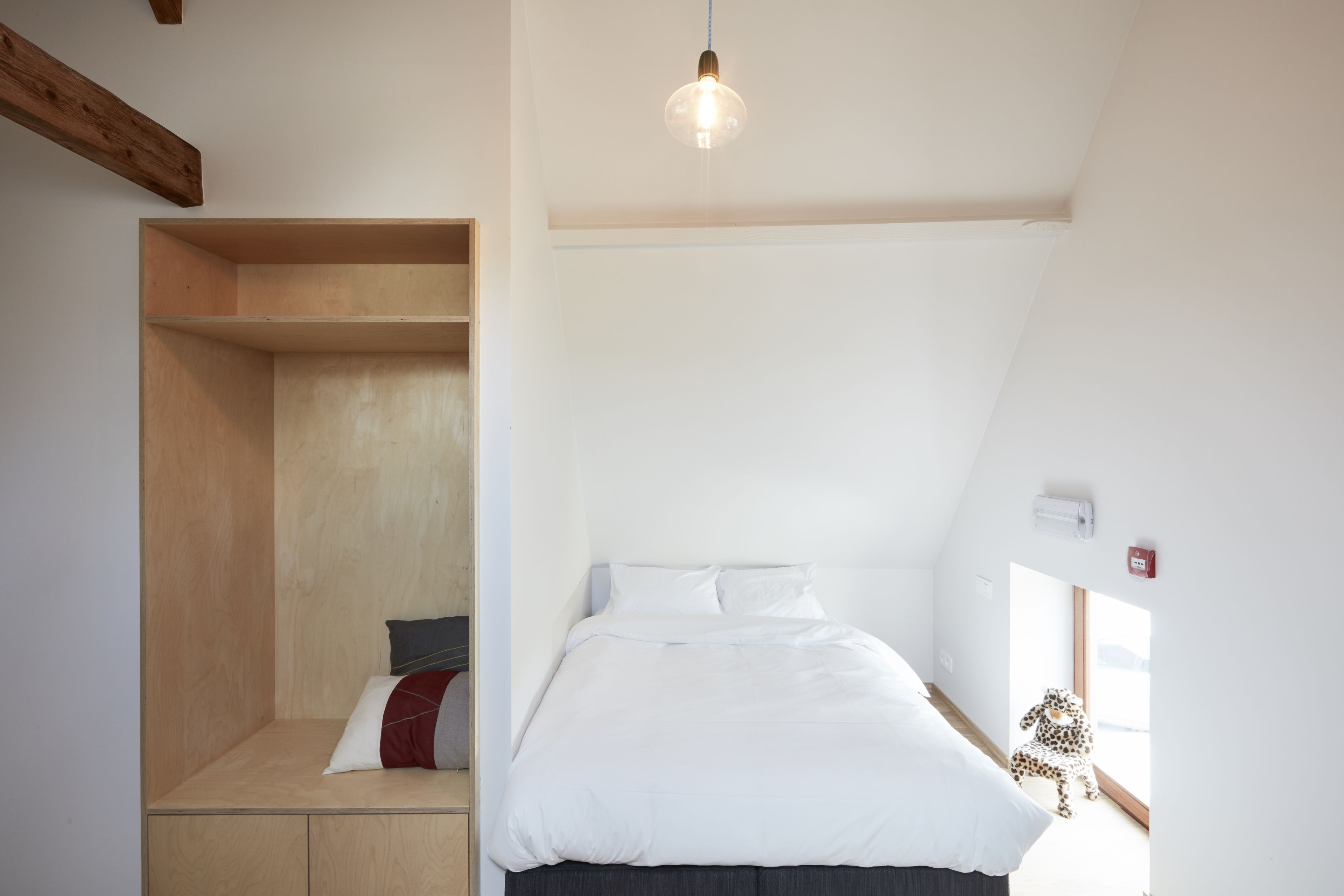 Een slaapkamer met een schuine kap, een bed met wit beddengoed, een houten bankje en een hanglamp.
