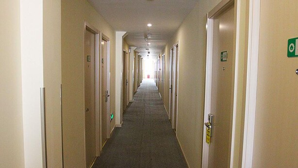 Un long couloir étroit, typique des hôtels, avec des portes des deux côtés, un sol recouvert de moquette et un plafond doté d’un éclairage encastré.