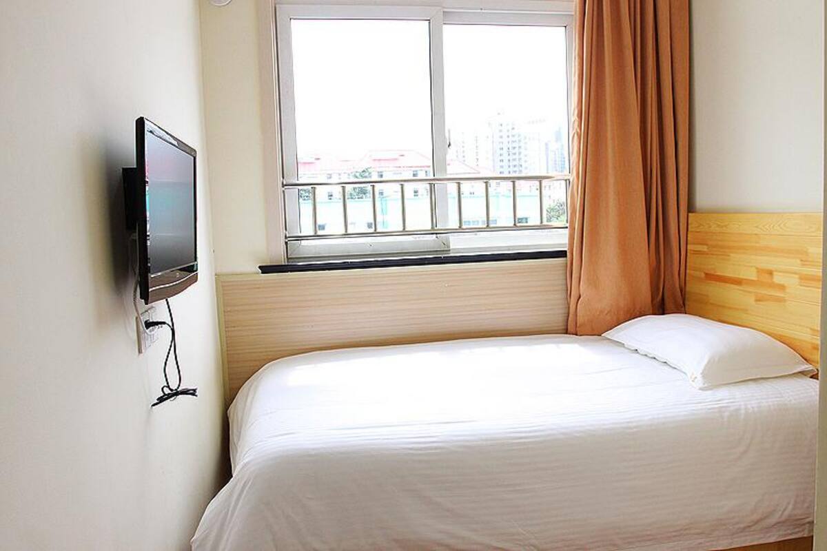 A hotel room with a bed, a TV mounted on the wall, a window with a view of buildings, and a curtain.
