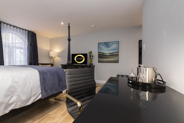 A modern hotel room with a bed, a desk with a tray of glasses, a lamp, a picture on the wall, and a window with curtains.