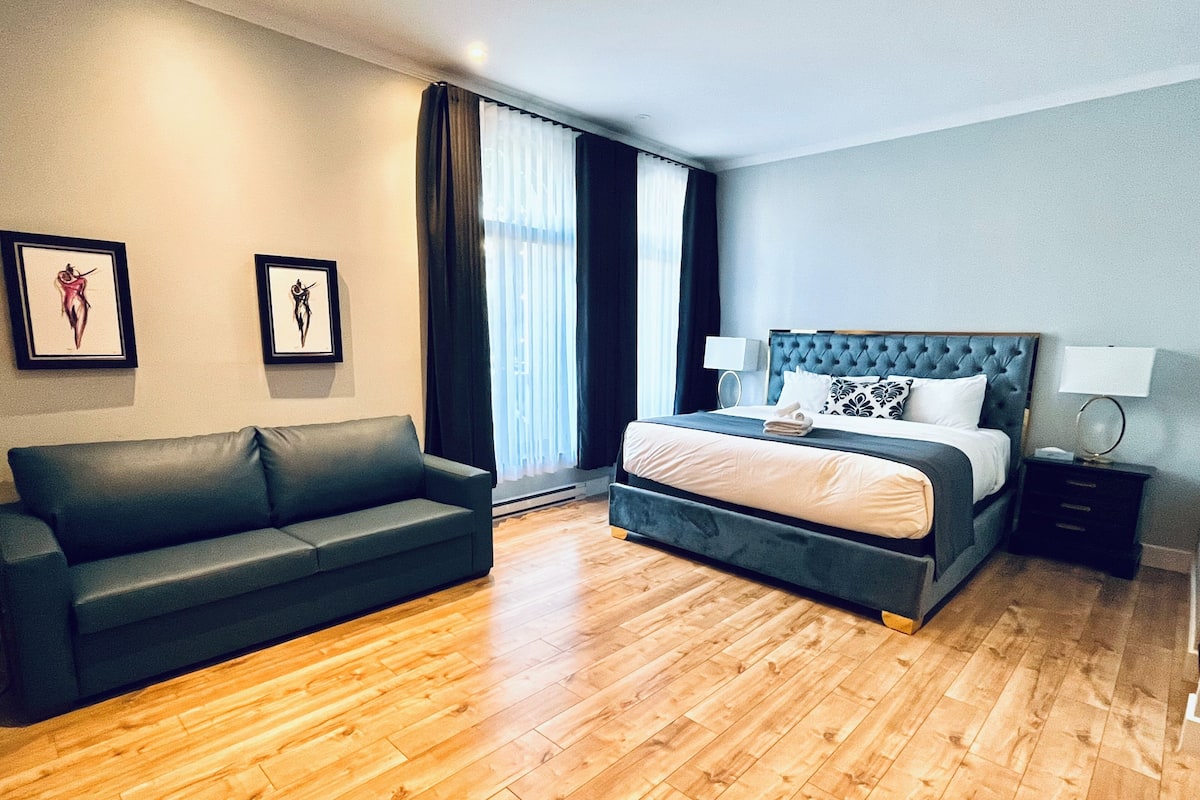 A modern hotel room with a large bed, a dark-colored sofa, and a wooden floor.