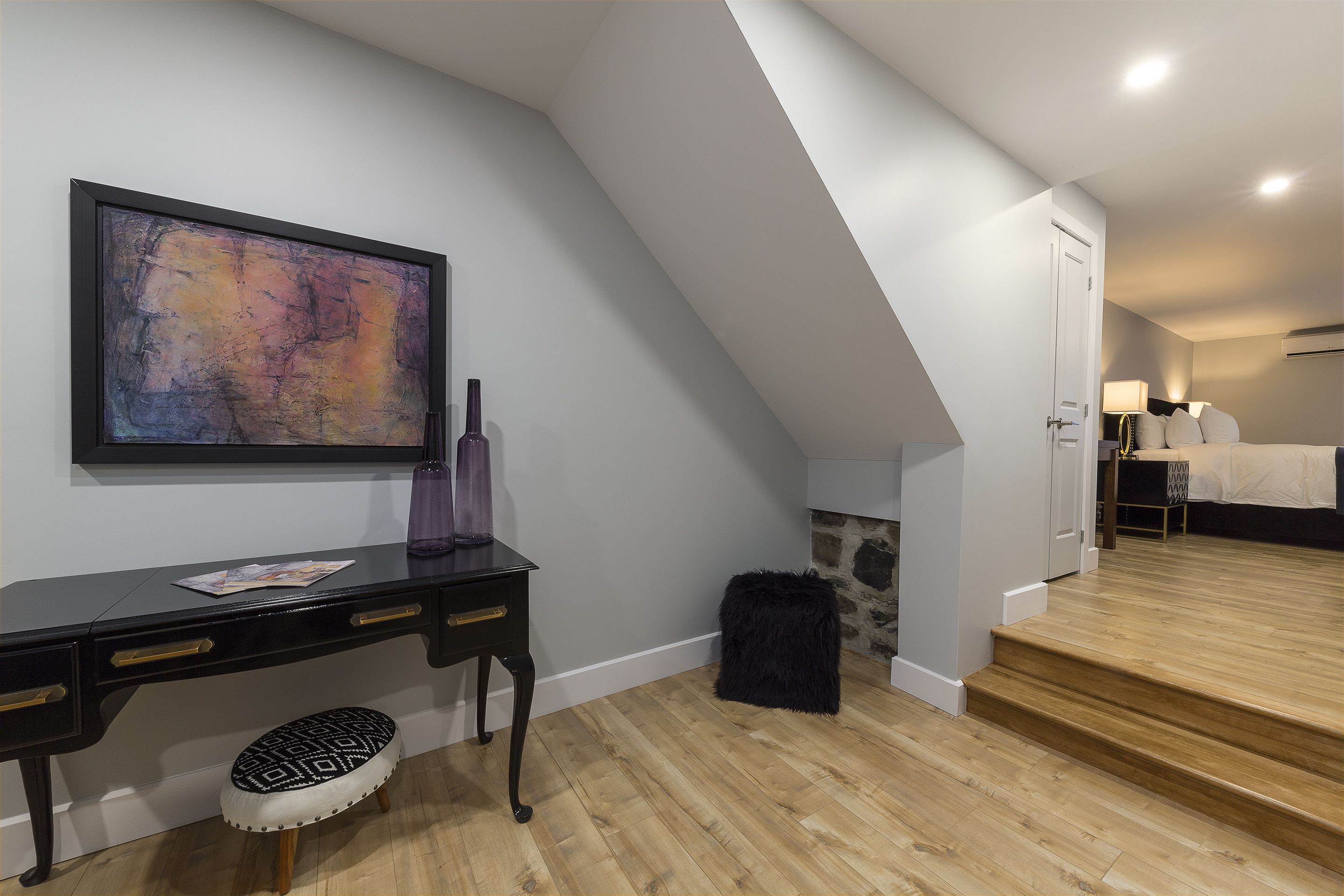 A modern bedroom with a wooden floor, a black desk with drawers, a black ottoman, a large abstract painting, and a staircase leading to another room.