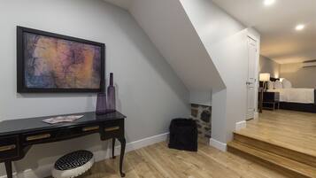 A modern bedroom with a wooden floor, a black desk with drawers, a black ottoman, a large abstract painting, and a staircase leading to another room.