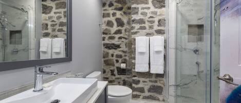 Une salle de bain avec un lavabo blanc, des toilettes, une douche à parois de verre et un mur en pierre.