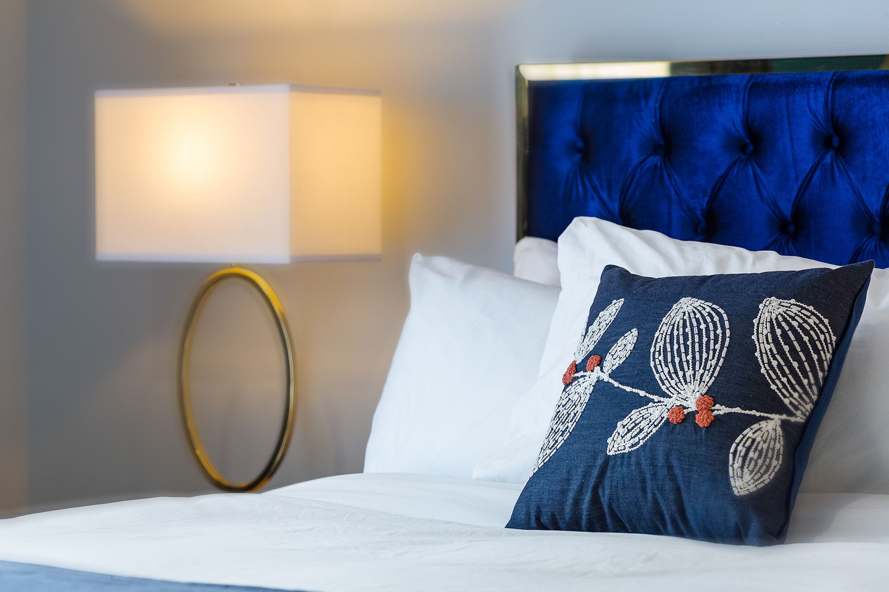 A neatly made bed with white linens, a blue upholstered headboard, and a decorative pillow with a floral pattern.