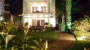 A two-story building with a balcony, surrounded by greenery and a paved pathway.