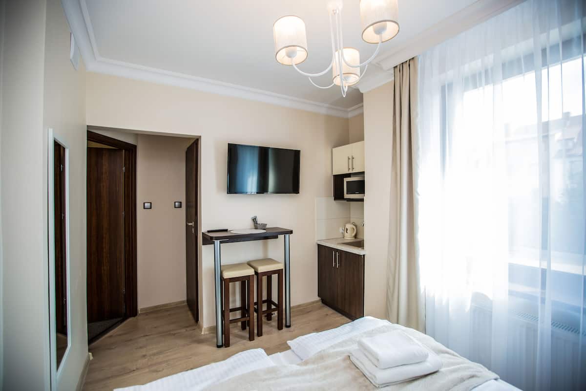 Ein Hotelzimmer mit einem Bett, einem Schreibtisch mit zwei Hocker, einem Fernseher, einer Küchenzeile und einem großen Fenster mit Vorhängen.