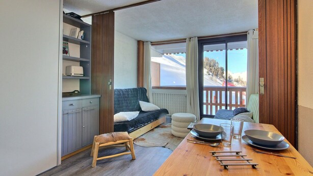 A compact room with a bed, a small stool, a dining table with cutlery, and a view of a snowy landscape through a sliding door. - Cozy studio with balcony and Mont-Blanc view in La Plagne (La Plagne)