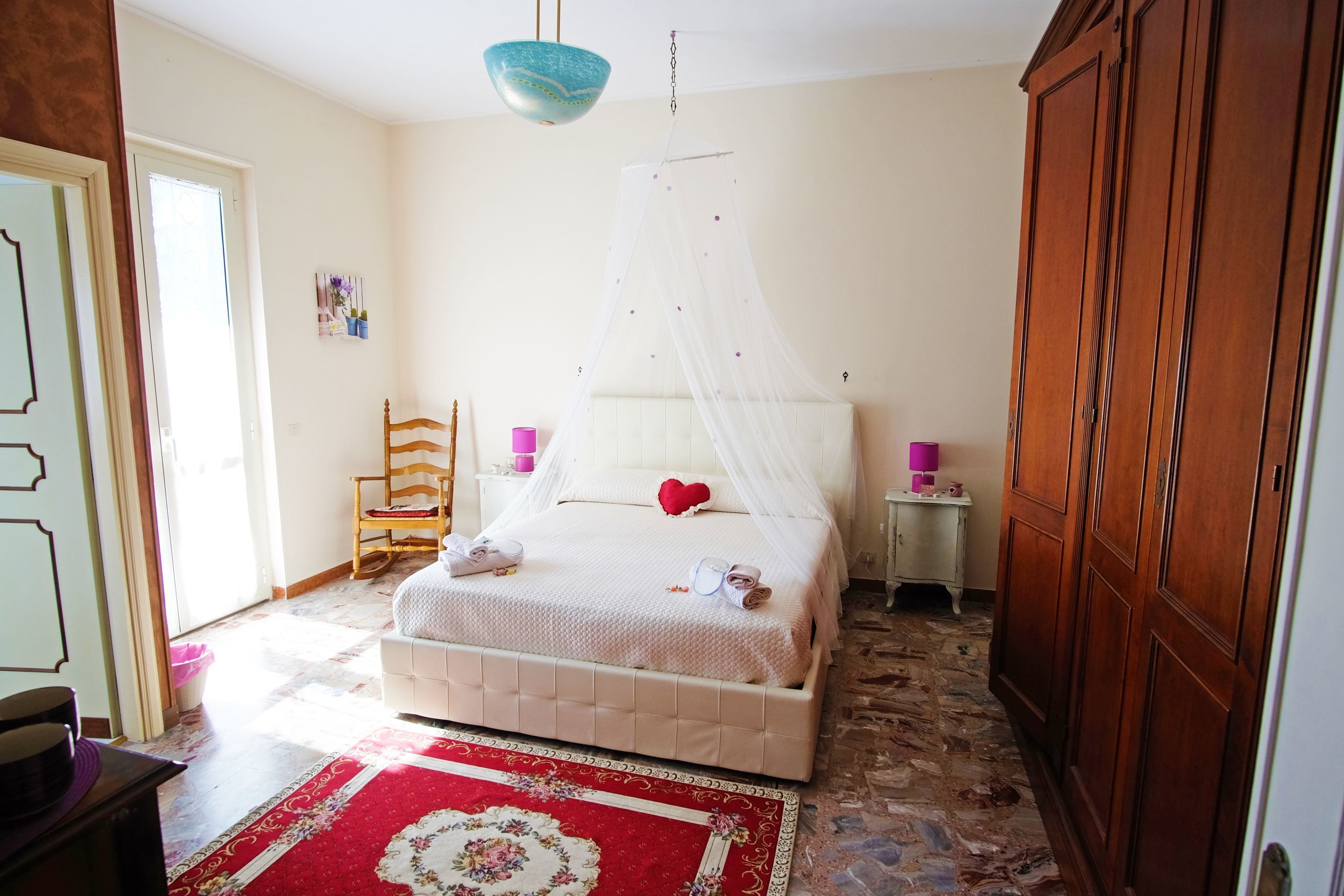 Une chambre à coucher comprenant un lit, une armoire en bois, une table de chevet, une chaise et un tapis à motifs rouges et blancs.