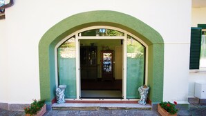 Une entrée en arc surmontée d’une porte en verre, flanquée de deux plantes en pot et ornée d’une statue en pierre sur les marches.