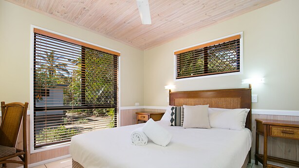 Una cama bien hecha con cabecera de madera, una silla y una mesita de noche con lámpara.