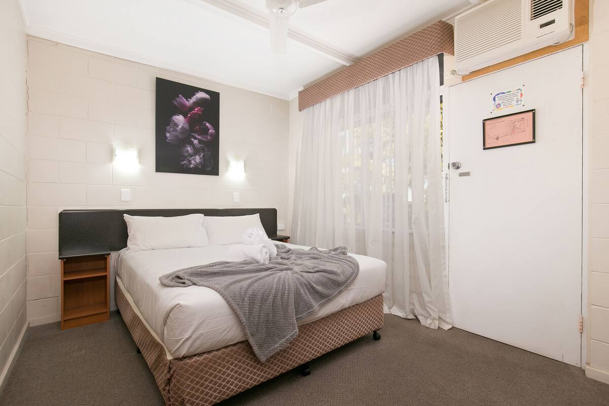 A neatly arranged bedroom with a bed, a nightstand, a wall-mounted light, a framed picture, and a door with a notice.
