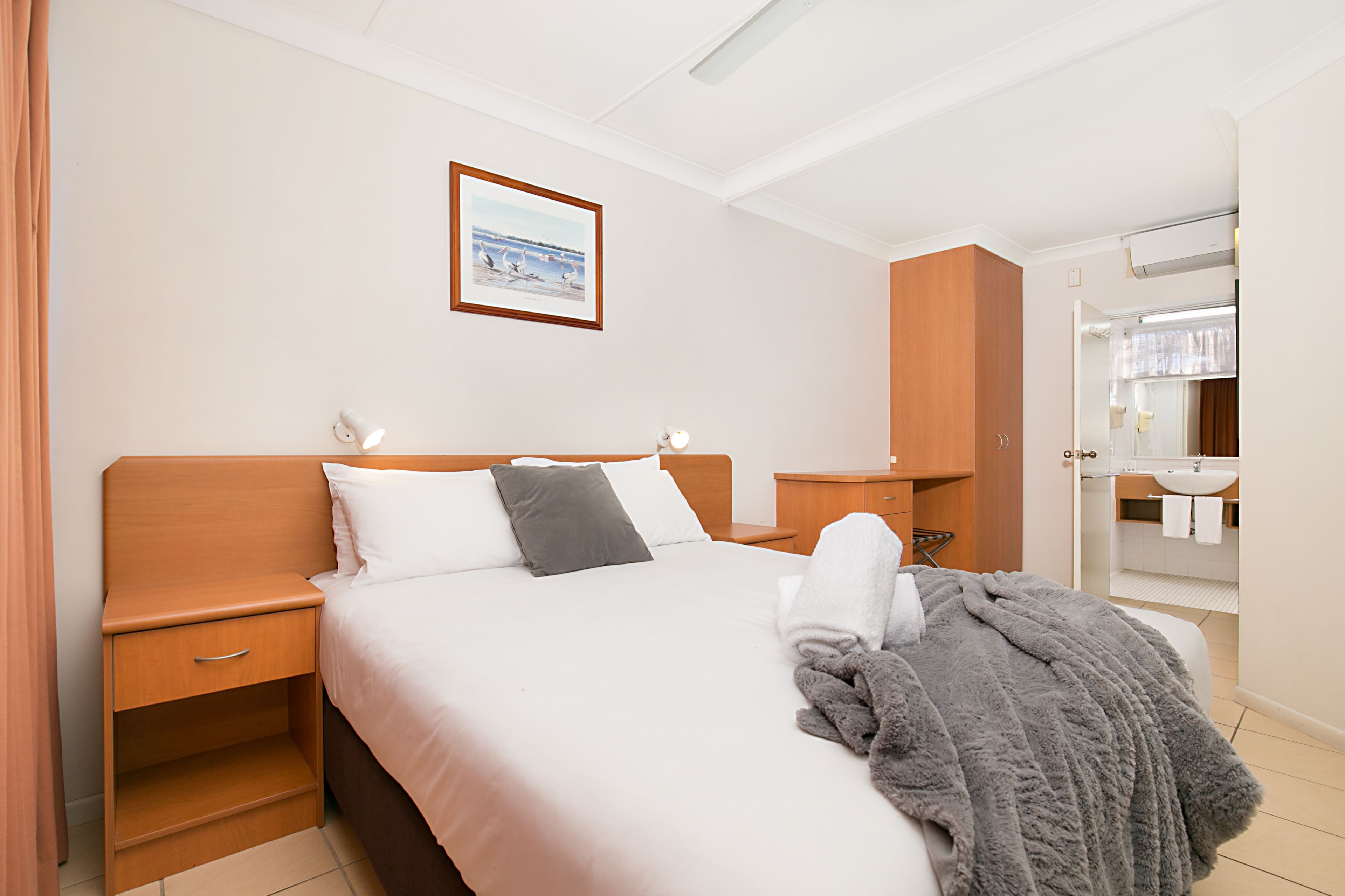 A hotel room with a bed, a nightstand, a wooden wardrobe, a bathroom with a sink and mirror.