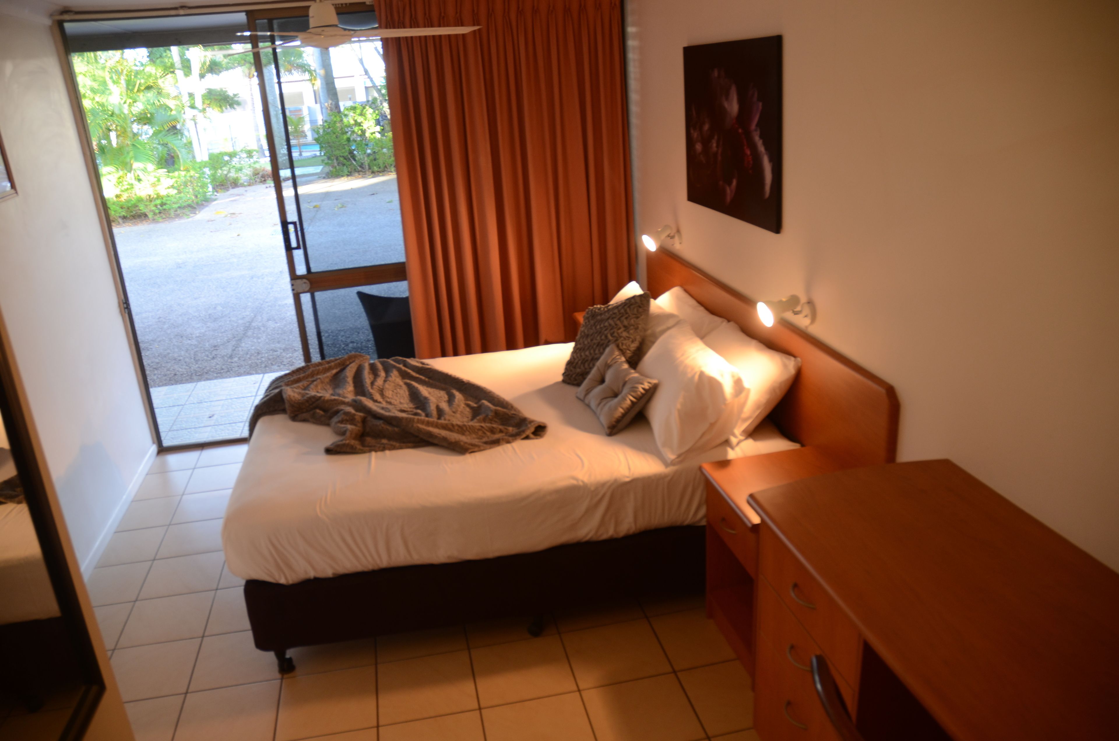 A hotel room with a bed, a wooden nightstand, a desk, and a view of the outdoors through a sliding door.