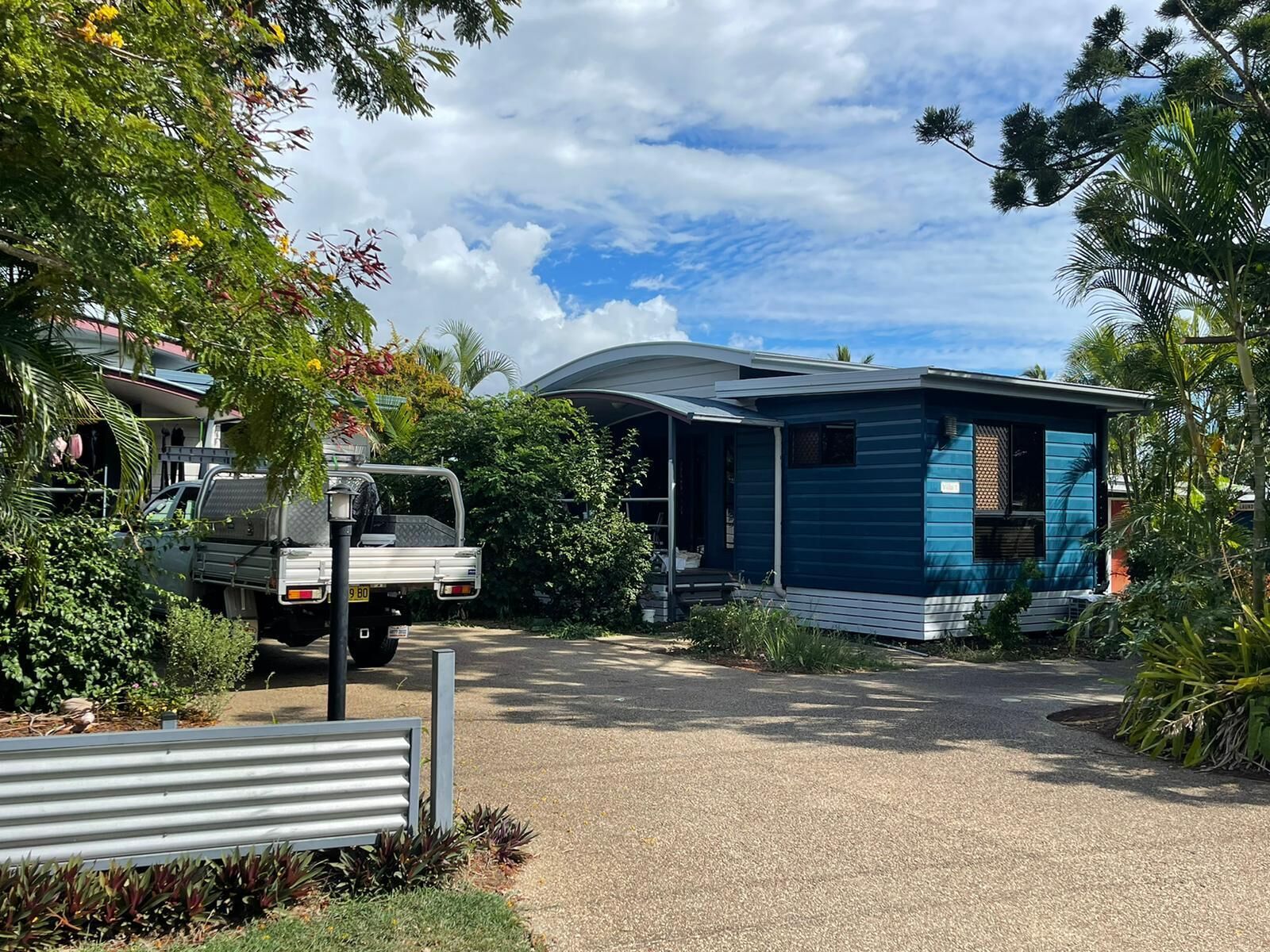 A mobile home with a blue exterior, a gravel driveway, and surrounding greenery.