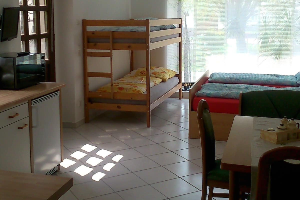 A room with a bunk bed, a desk with a computer, a chair, and a table with a teapot.