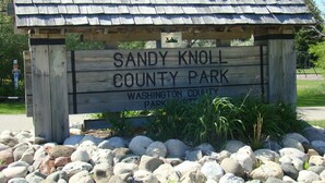 Un panneau en bois indiquant le parc du comté de Sandy Knoll, surmonté d’un toit en métal, et entouré de rochers et de verdure.