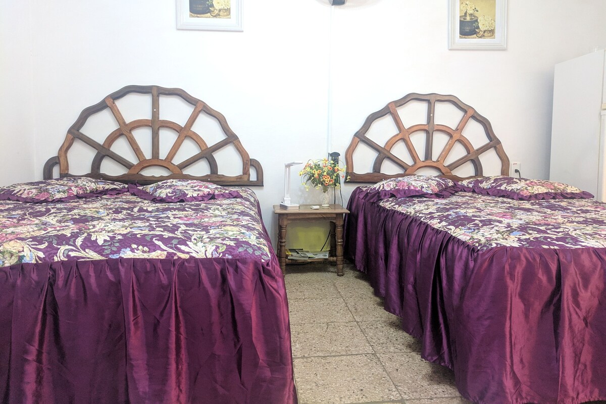 Ein Zimmer mit zwei Betten, jeweils mit einem hölzernen Kopfteil und violetter Bettwäsche. Dazwischen steht ein Nachttisch mit einem Blumenstrauß.