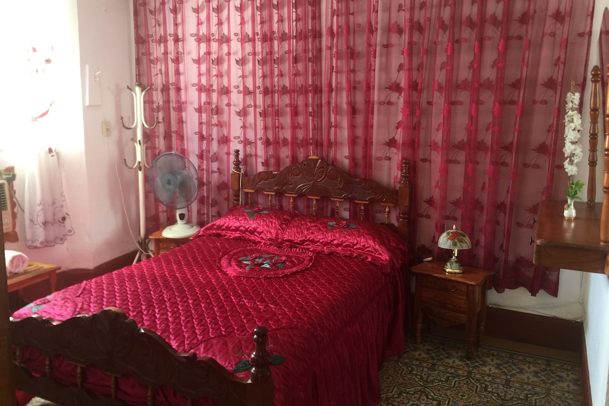 Une chambre à coucher avec un lit en bois, une table de chevet avec une lampe, un ventilateur et un rideau à motifs floraux.