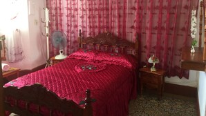 A bedroom with a wooden bed, a bedside table with a lamp, a fan, and a floral curtain. - Casa Colonial Center (Cienfuegos)