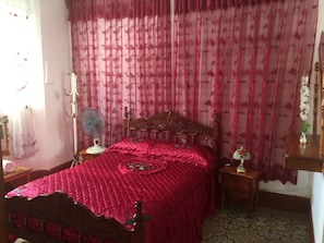 Une chambre à coucher avec un lit en bois, une table de chevet avec une lampe, un ventilateur et un rideau à motifs floraux.