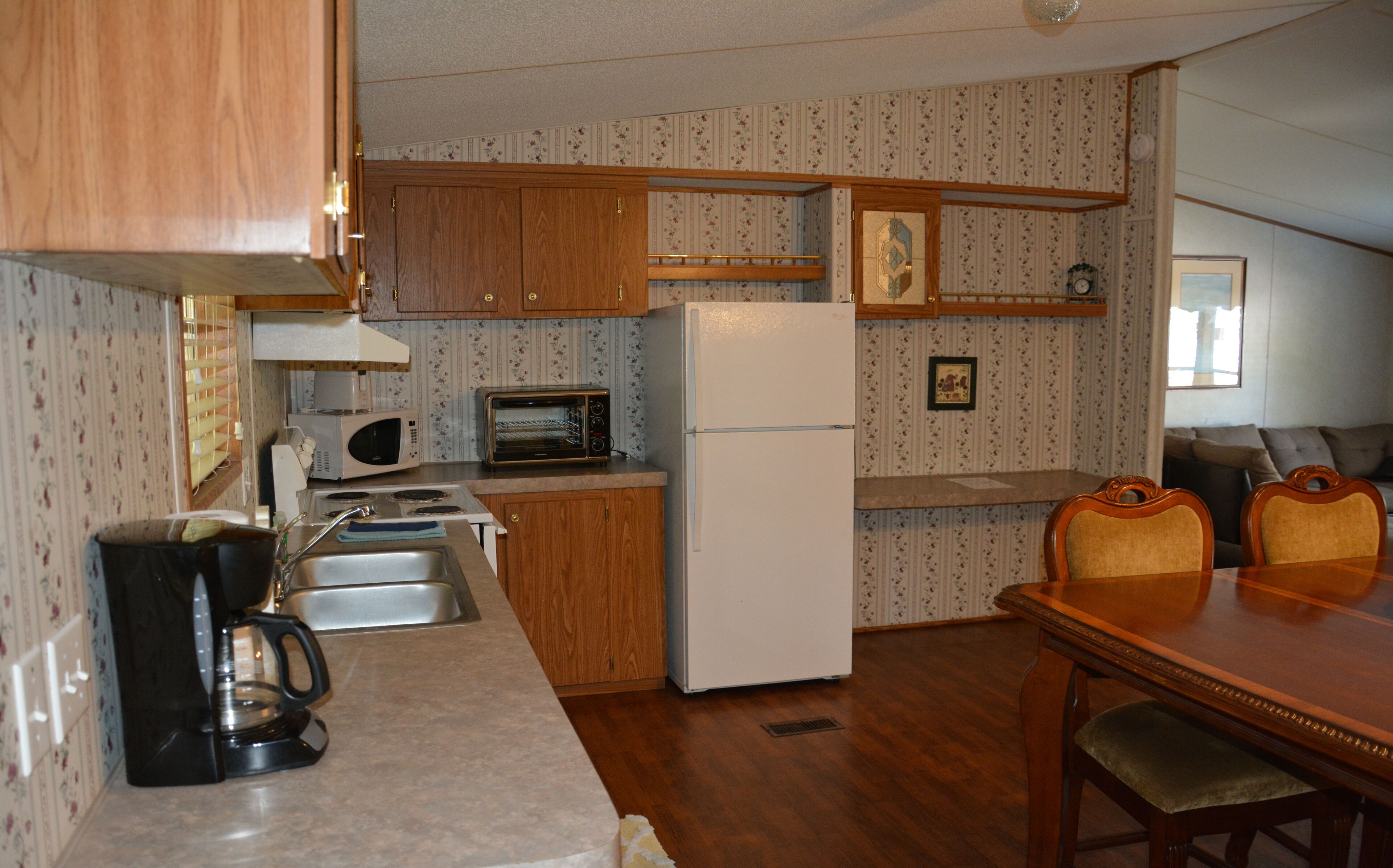 A compact kitchen with wooden cabinets, a white refrigerator, a microwave, and a toaster. There is a dining table with chairs and a small window.