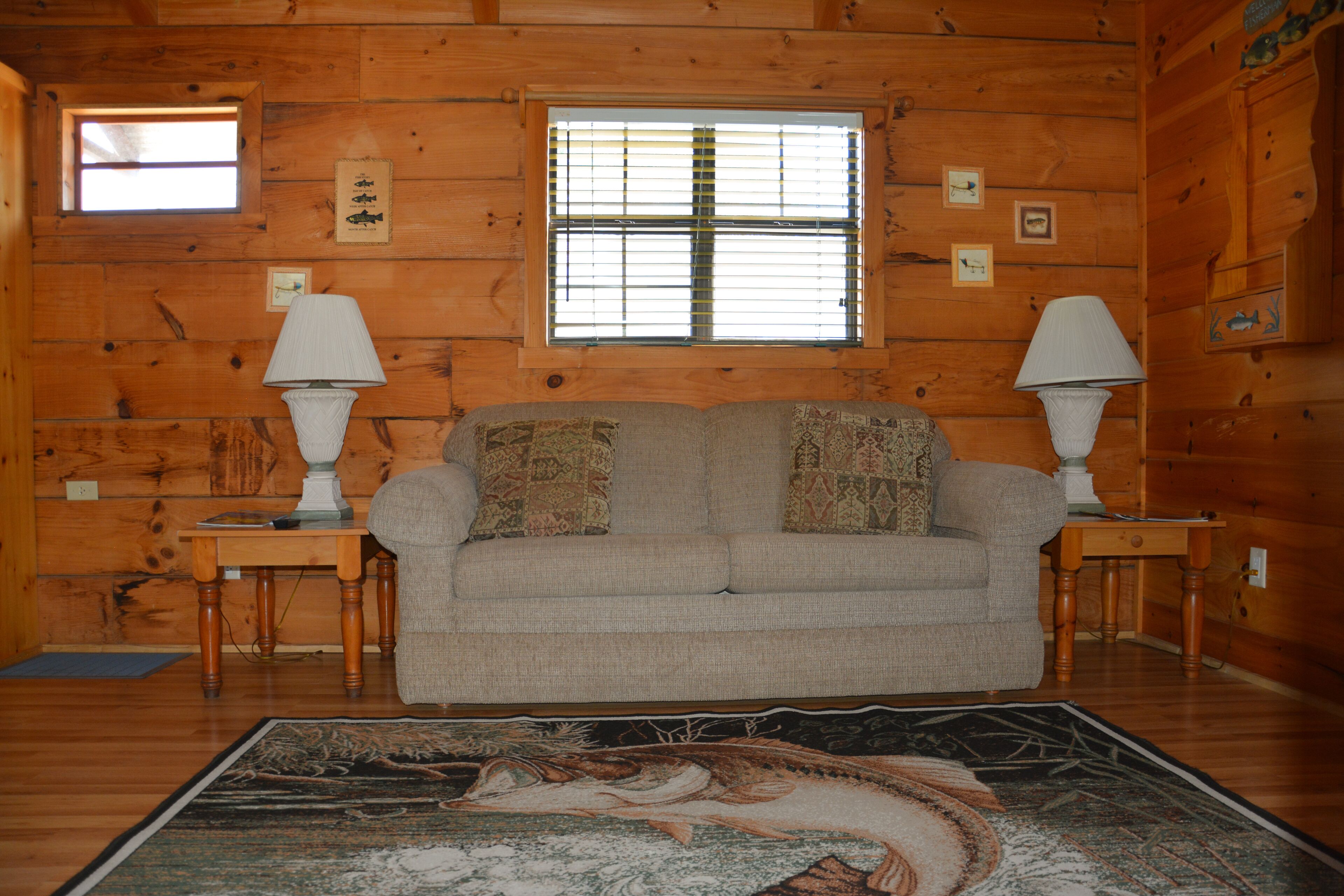 A cozy log cabin interior with a beige sofa, two side tables with lamps, a large window with blinds, and a decorative rug.