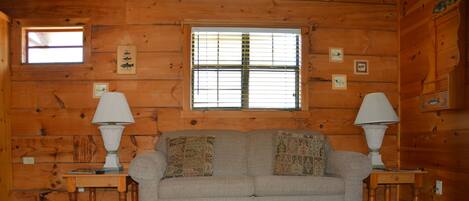 A cozy log cabin interior with a beige sofa, two side tables with lamps, a large window with blinds, and a decorative rug.