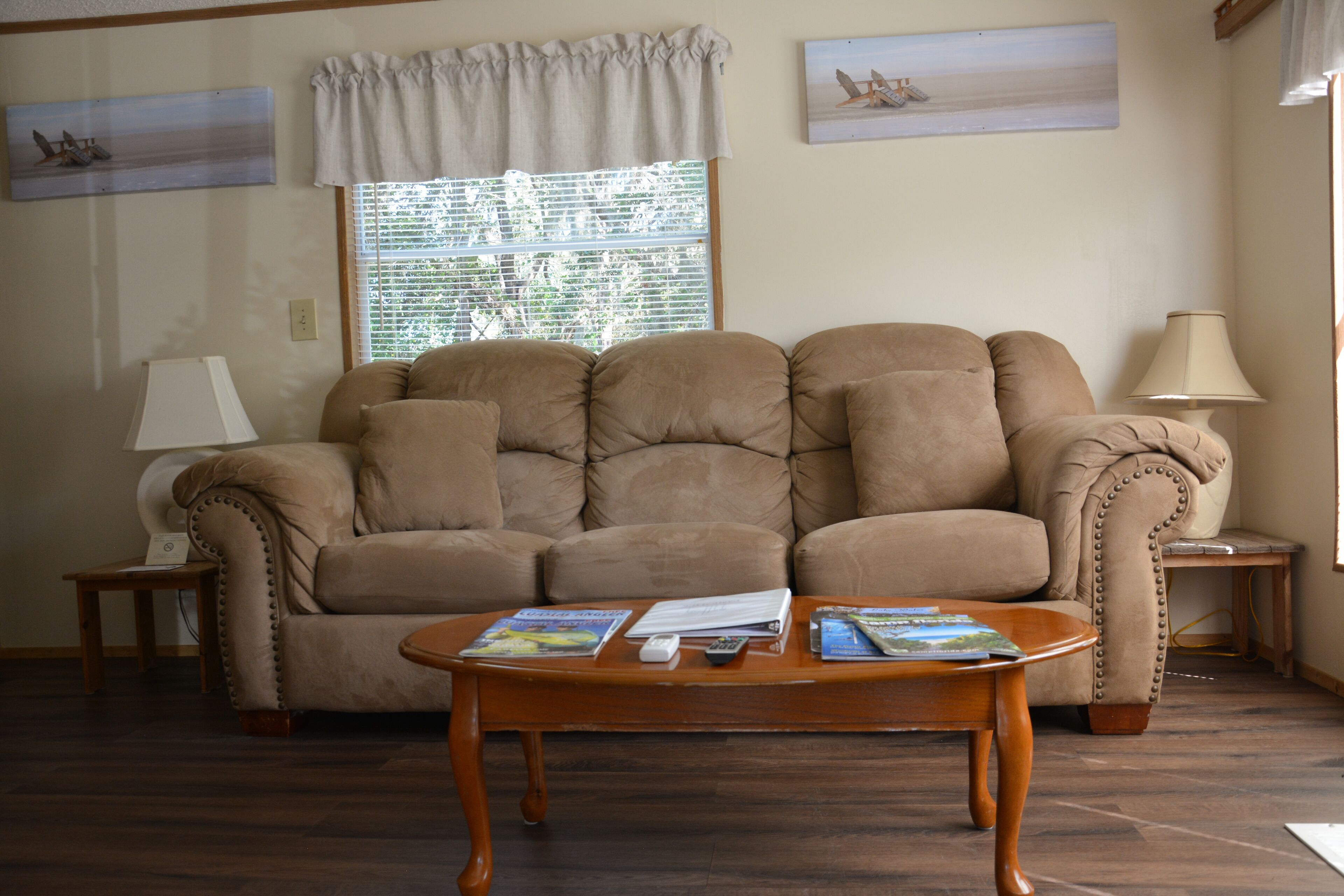 A living room with a beige sofa, a wooden coffee table with magazines, a lamp, and a window with blinds.