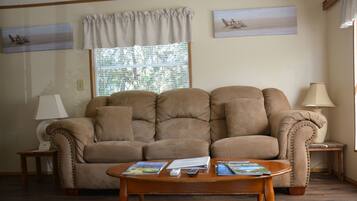 A living room with a beige sofa, a wooden coffee table with magazines, a lamp, and a window with blinds.