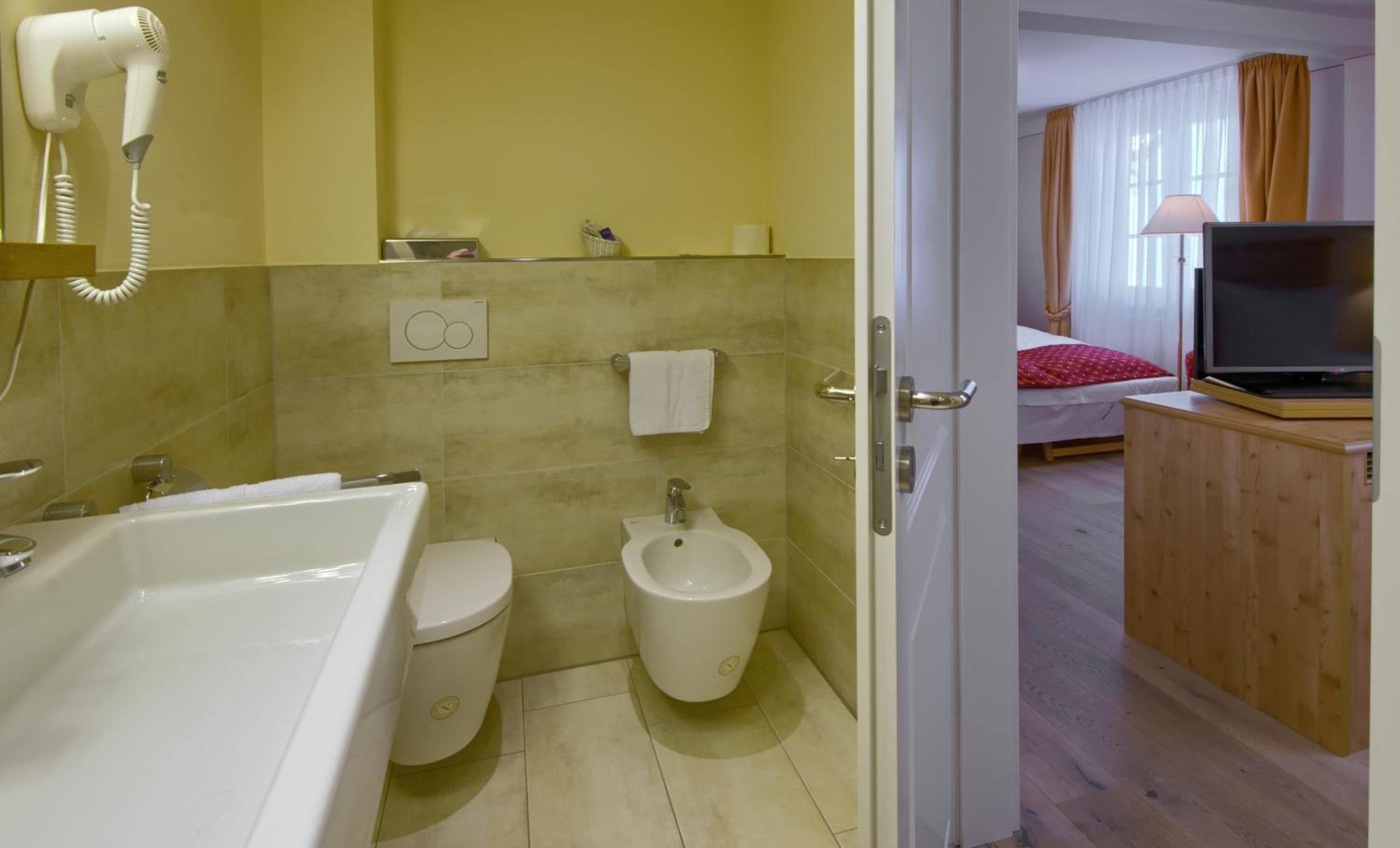A bathroom with a bathtub, toilet, and bidet. There is a hairdryer on the wall and a towel hanging on the wall.