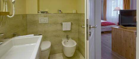 Een badkamer met een bad, toilet en bidet. Er is een haardroger aan de muur en een handdoek hangt aan de wand.
