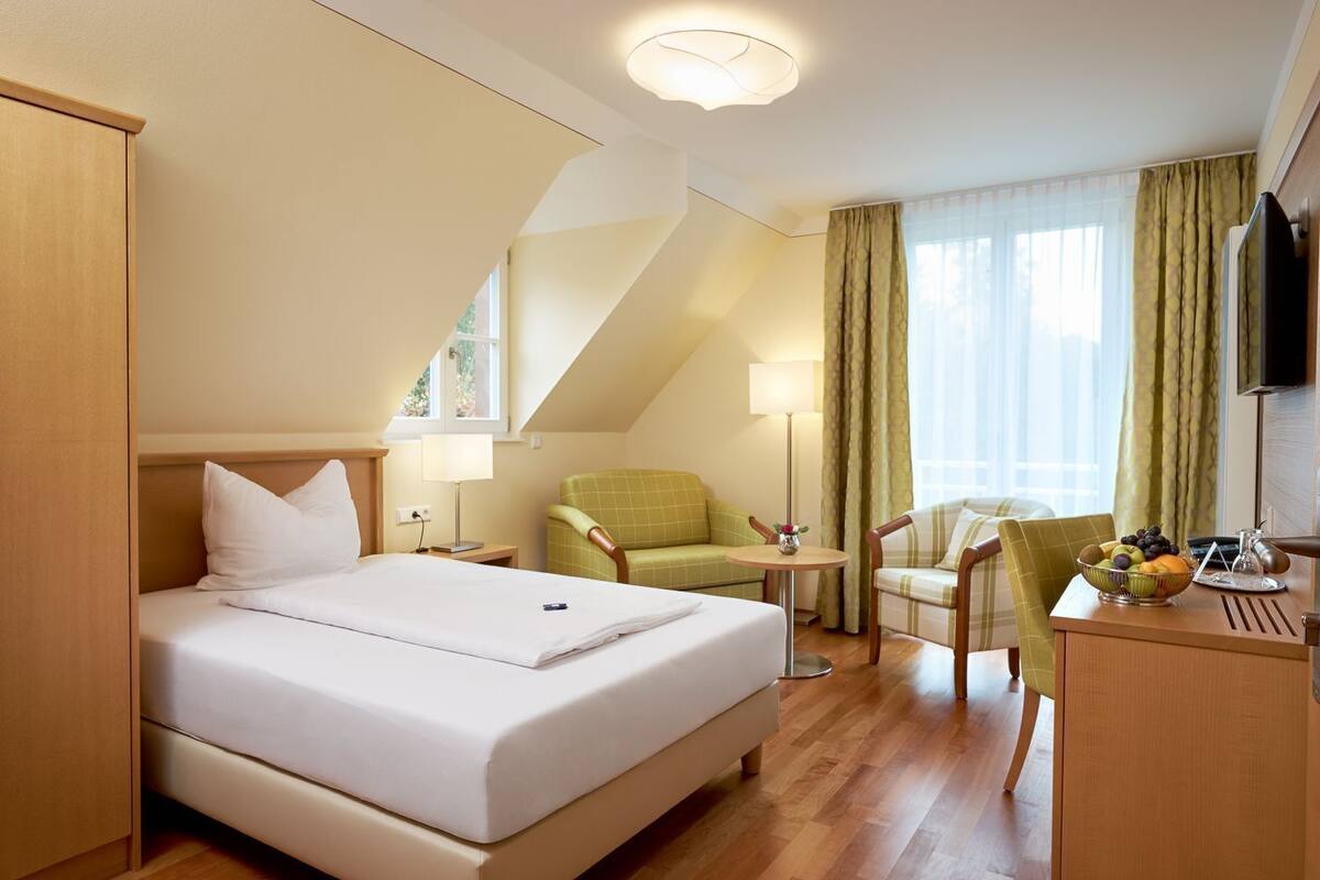 A hotel room with a bed, two armchairs, a desk with a fruit bowl, a lamp, and a TV.