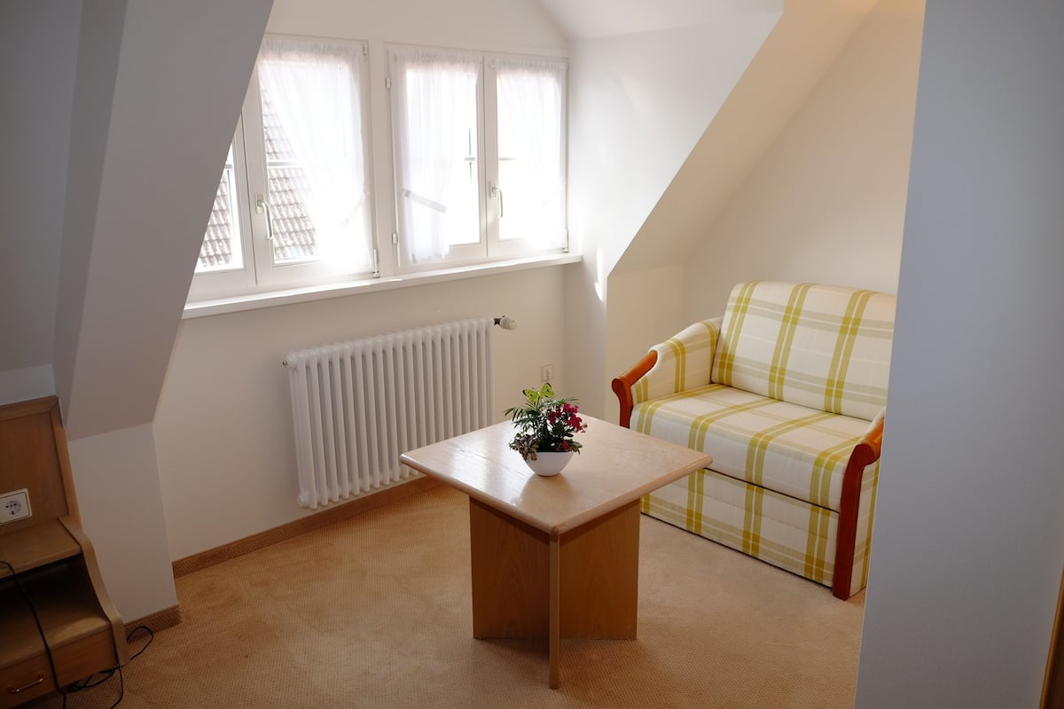 A small, well-lit room with a sloped ceiling, a radiator, a wooden table with a flower pot, and a plaid sofa.