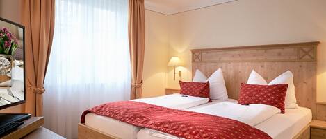 A hotel room with a wooden bed, beige curtains, a bedside lamp, and a desk with a computer.