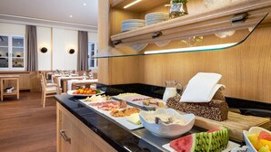 A buffet with various food items, a seating area with tables and chairs, and a wooden wall with shelves holding plates and glasses. - Flair Hotel Schwanen (Bad Bellingen)