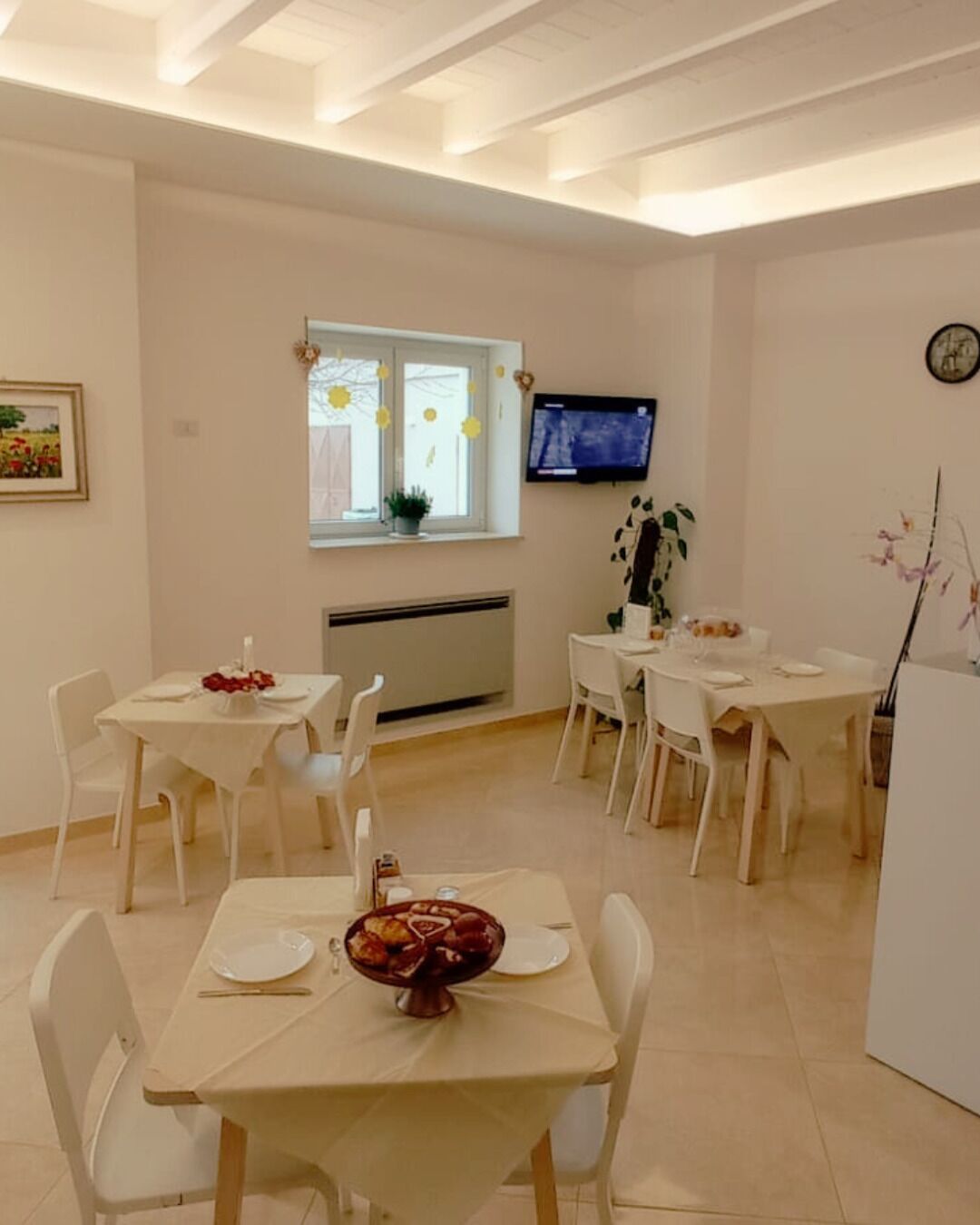 A dining area with white tables and chairs, a TV mounted on the wall, and a window with decorations.