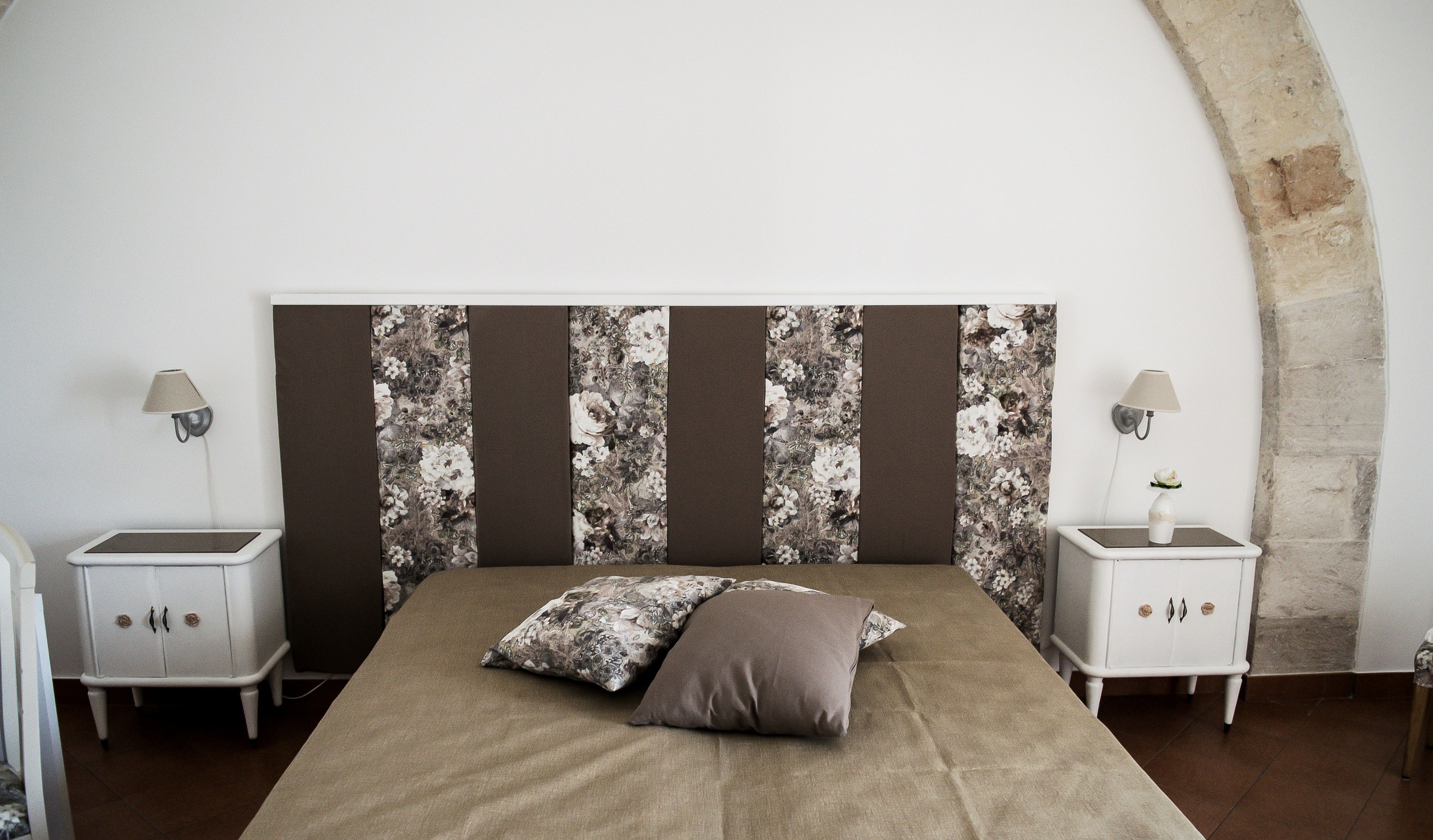 A bedroom with a large bed, two nightstands, a headboard with a floral pattern, and a stone archway.