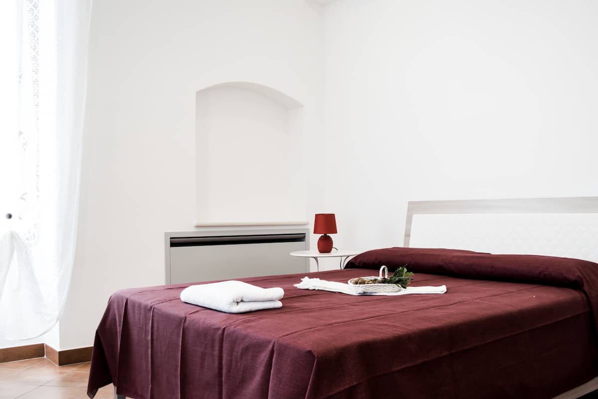 Ein ordentlich bezogenes Bett mit roter Tagesdecke, einem weißen Kissen und einem weißen Handtuch auf der Seite. Daneben steht ein kleiner Tisch mit einer roten Lampe und einem dekorativen Gegenstand.