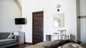 A modern room with a dark wooden door, a flat-screen TV mounted on the wall, a white chair, a small table, and a sofa with cushions. - Tenuta Vigna Del Noce (Corato)