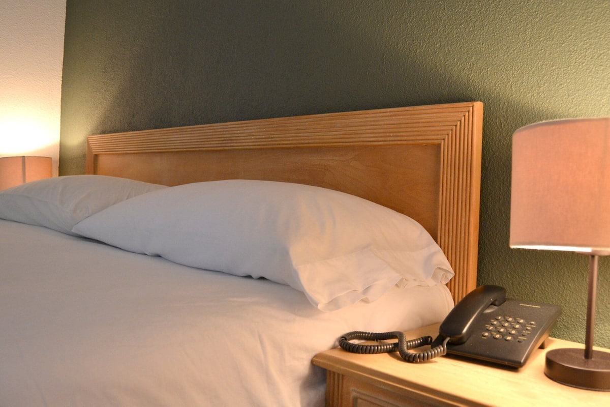 Un letto rifatto con lenzuola bianche, una testiera in legno, un telefono sul comodino e una lampada su un poggiabibite in legno.